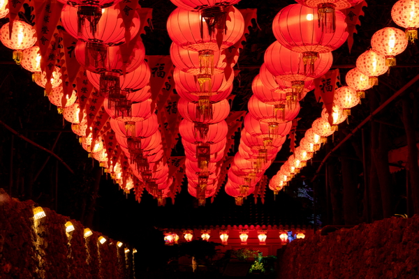 暗闇の中に、赤い丸型のランタンが何列にもわたって奥行きを持って吊るされている様子。提灯の間には赤い旗が飾られ、下方には石垣がライトアップされている。