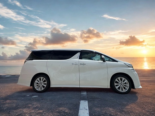 美しい夕日と海を背景に、海岸沿いに停車しているMOKU・MOKUレンタカーの白い新型ヴェルファイアの真横(サイドビュー)のカット。