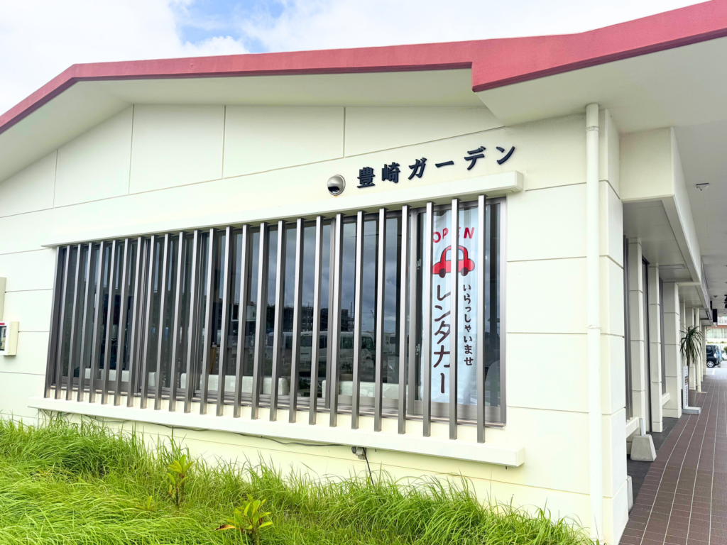白い壁と赤フチの屋根が特徴的な「豊崎ガーデン」の建物外観とレンタカーの案内旗